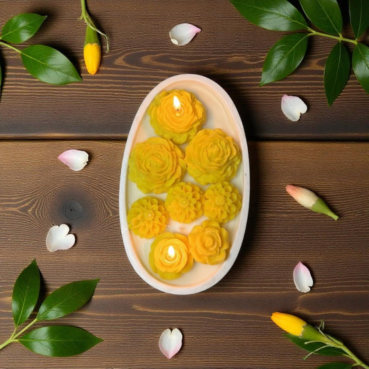 Top-down view of an oval white ceramic dish candle featuring bright yellow, detailed floral wax embeds, lit with two small flames, set against a dark rustic wood background.