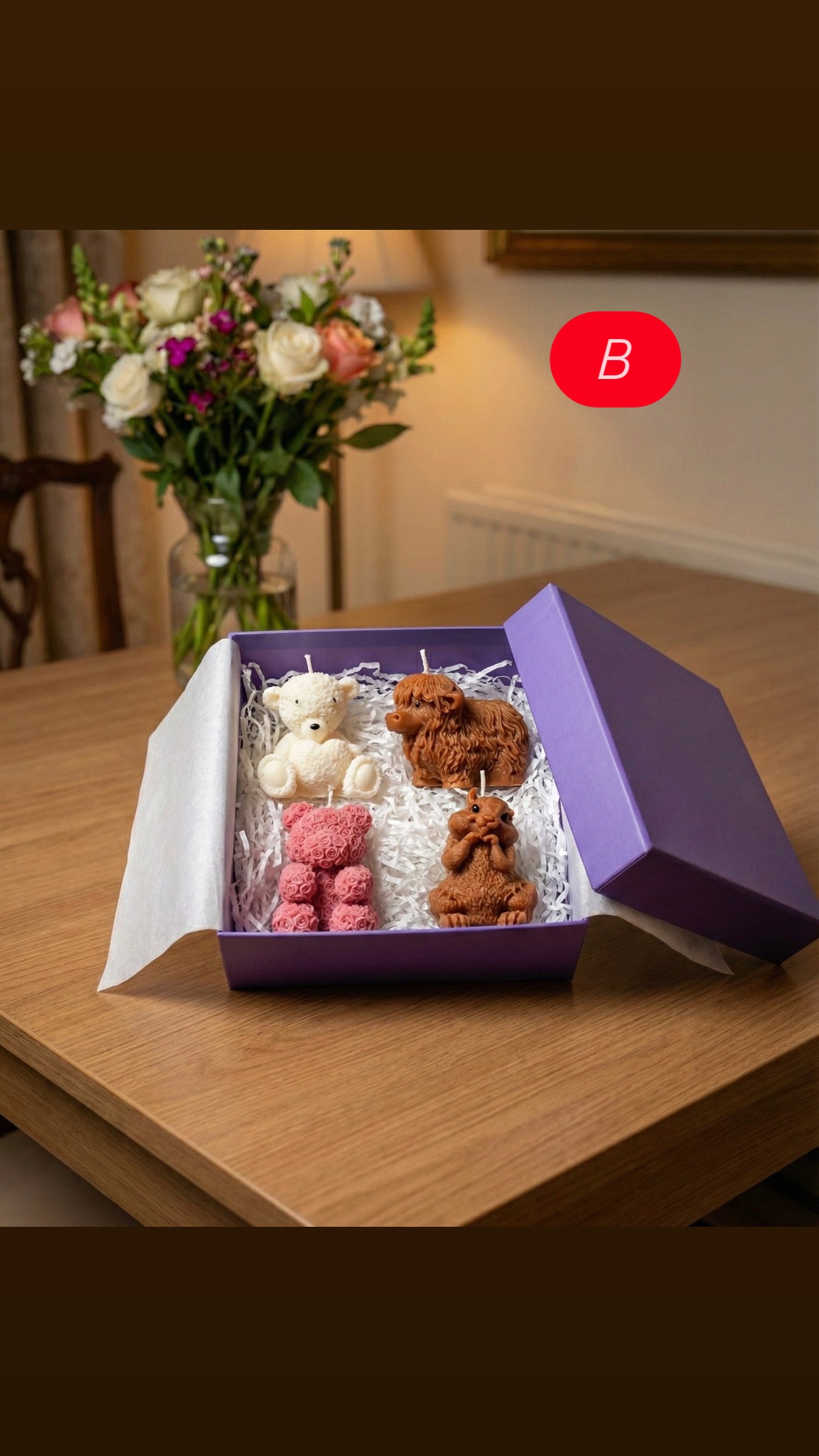 Open purple gift box labeled B sitting on a wooden table. The box contains four shaped candles: a white sitting teddy bear, a brown highland cow, a pink rose-textured bear, and a brown squirrel holding a nut.