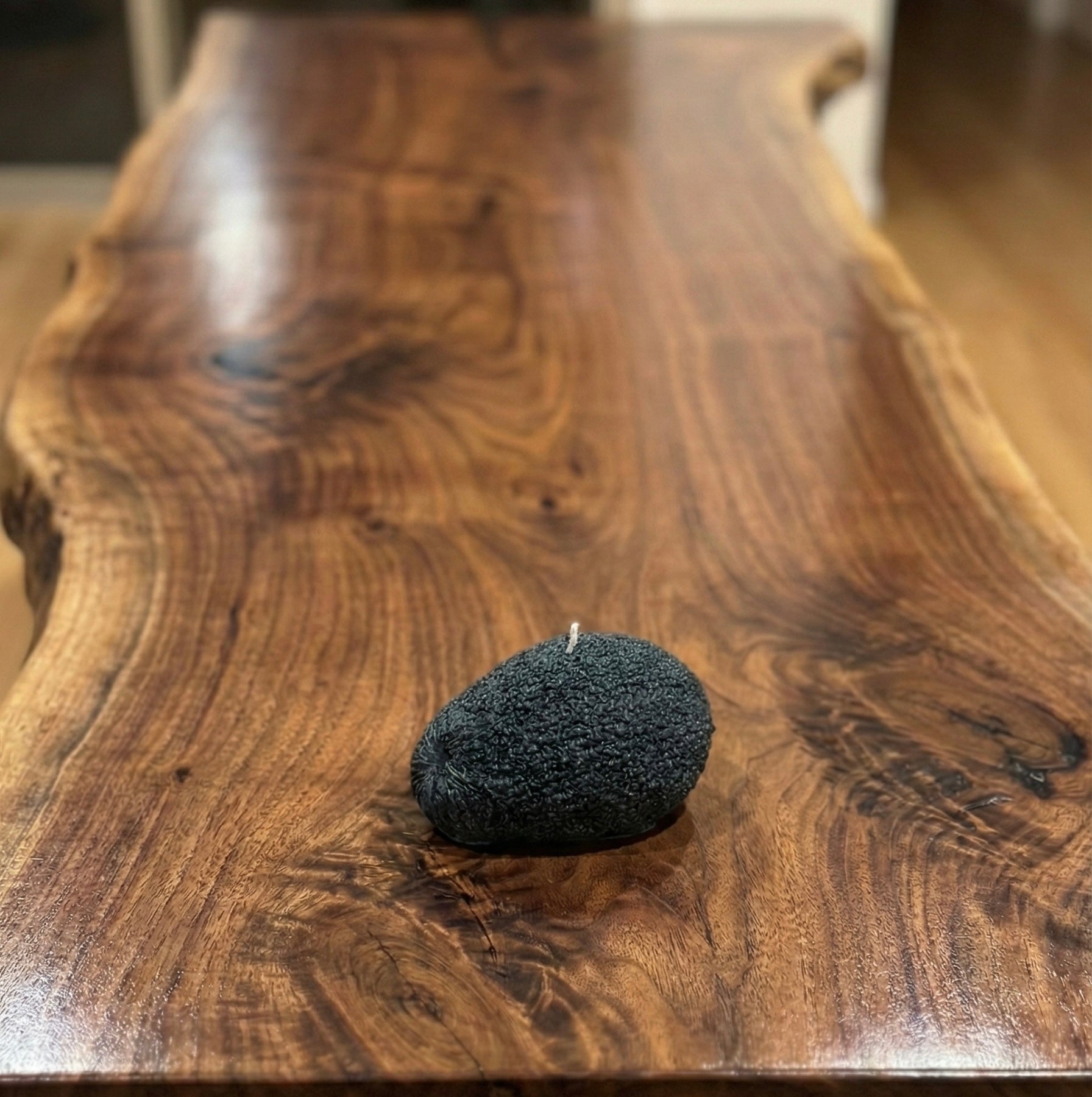 A hyper-realistic candle shaped like a whole avocado with a textured, bumpy skin surface in a deep black charcoal color, sitting on a polished live-edge walnut wood table. The candle has a single white wick at the top."