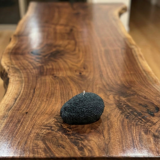 A hyper-realistic candle shaped like a whole avocado with a textured, bumpy skin surface in a deep black charcoal color, sitting on a polished live-edge walnut wood table. The candle has a single white wick at the top."