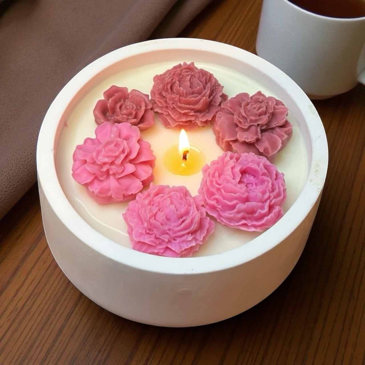 Lit Round White Bowl decorative candle featuring a ring of pink peony-style wax flowers. Peony centerpiece styled with a coffee mug and fabric. Luxurious floral soy wax."